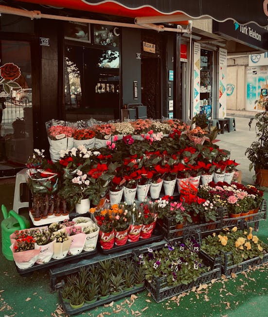 A colorful display of fresh flowers arranged on black plastic tiered racks outside a flower shop. The arrangement features a variety of blooms including red roses, pink carnations, white and purple pa