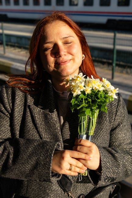 A woman with long red hair and closed eyes is holding a bouquet of fresh, vibrant yellow and white daffodils with green foliage, wrapped loosely in clear cellophane. She is wearing a dark grey coat an