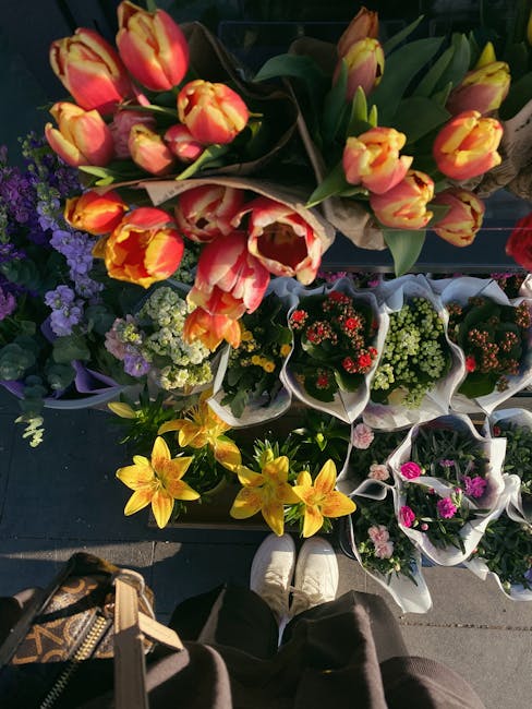 A vibrant display of freshly cut tulips with orange and yellow petals and green stems, arranged in a bouquet wrapped in brown paper, is held above an assortment of potted flowers on a market stall. Su