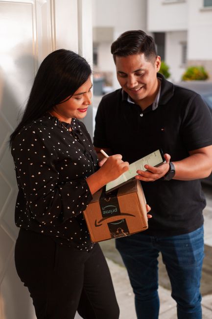 A woman with long dark hair and a black dress with white heart patterns is smiling as she signs a receipt on a clipboard held by a man with short dark hair, wearing a dark polo shirt. They are standin