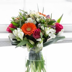 This stunning bouquet, available for delivery throughout the Acton community, is a study in harmonious colour and elegant structure, thoughtfully curated for both visual and emotional impact. Cream-hued gerbera daisies form a radiant outer ring, their silky petals gently framing the bold, zesty orange centres that catch the light like lanterns at dusk over Heathfield Terrace. Lush magenta roses are carefully nestled amidst this halo, their deeply coloured petals adding drama and a sense of romantic refinement. Scattered through the arrangement, pristine white lilies-possibly delicate alstroemerias-intermingle with small white bloom clusters, creating layers of airy brightness and enhancing the bouquet's garden-fresh feel. Supporting the florals is an emerald embrace of broad, glossy aspidistra leaves and verdant paddle-shaped greens, resting gracefully around the base to reinforce the bouquet's rounded silhouette and provide textural contrast. The clear vase reveals the sturdy tangle of natural stems, a tangible reminder of the flowers' freshness and longevity. Natural daylight pours over the arrangement, highlighting each scent and shade-a floral composition perfect for a celebratory brunch, marking a new beginning, or simply bringing the spirit of Acton's thriving markets into your home. Every carefully placed bloom reflects the expertise and creative flair characteristic of local florists here in W3.